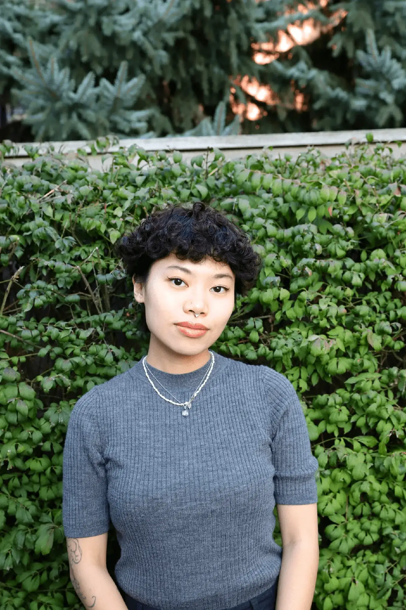 Anhi T, Academic and Portfolio Advisor, in a grey sweater in front of a green hedge