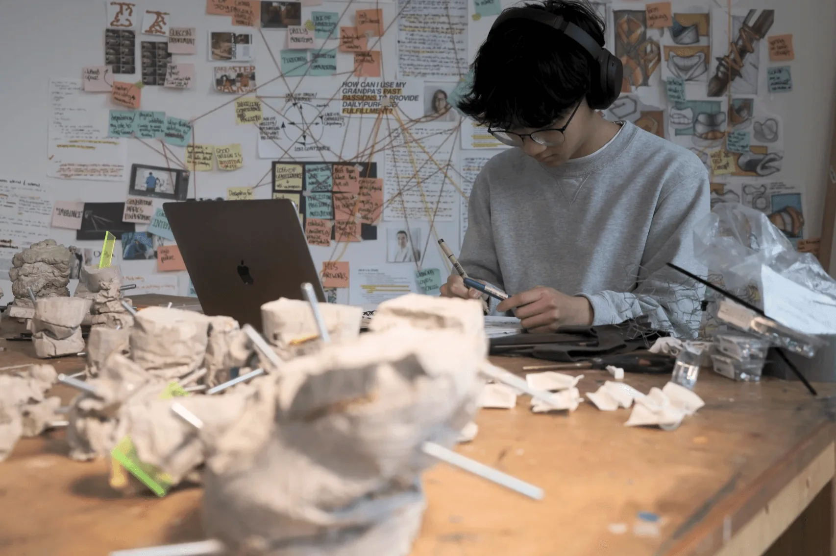 Student in headphones sculpting paper forms at a desk beneath a wall of research notes
